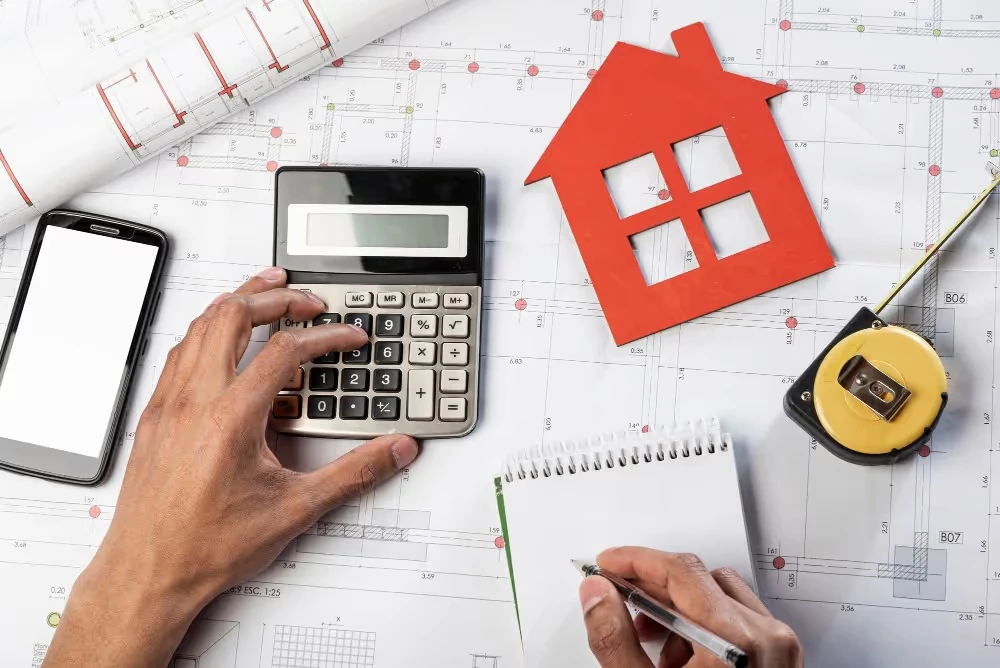 Calculatrice, silhouette de maison et mètre ruban sur un plan, illustration de la simulation d’un prêt immobilier.