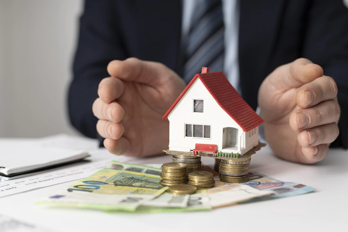 Des mains entourent une maquette de maison sur des billets et des pièces, illustration de l’investissement locatif sans apport.